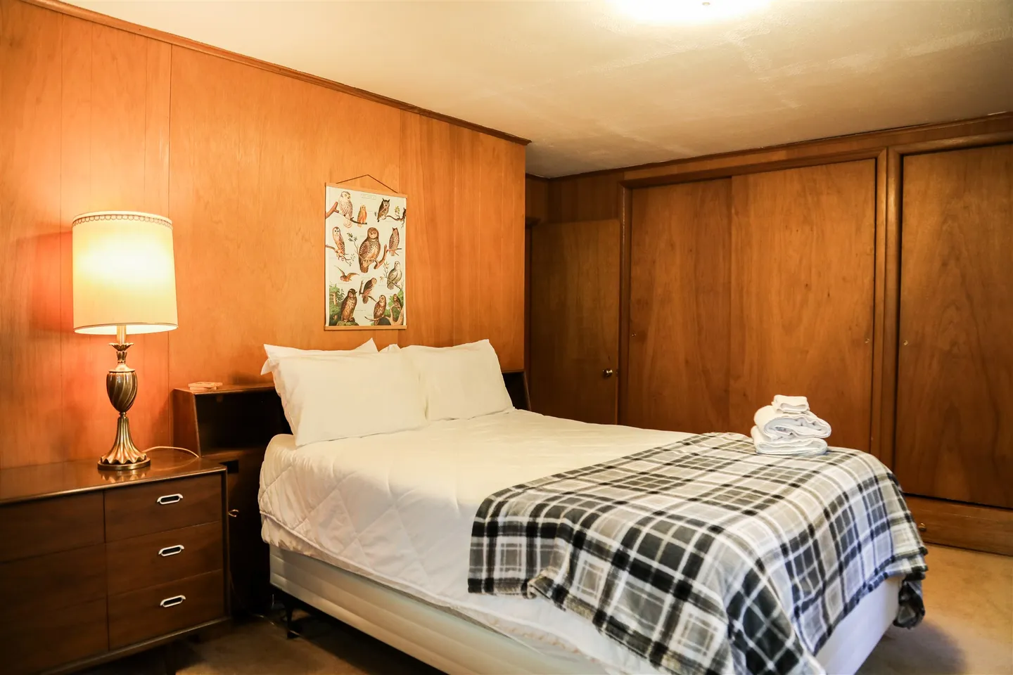 Bedroom featuring wood paneled walls, a made bed with a plaid throw blanket, a nightstand with a lamp, and sliding closet doors.
