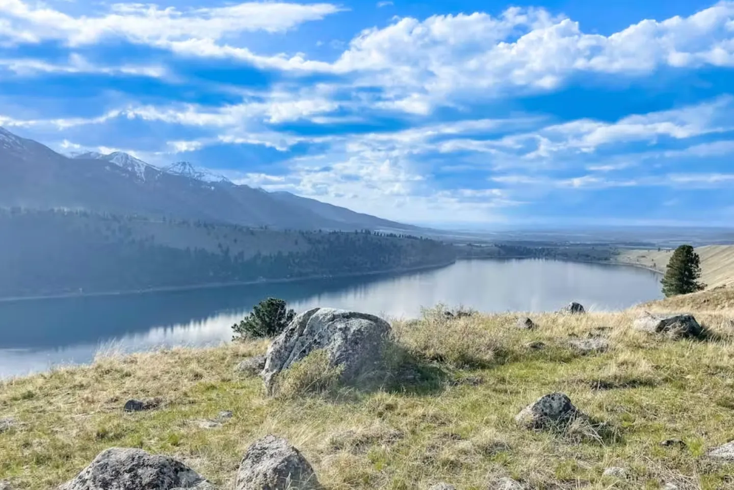 Elevated outdoor view overlooking a calm lake surrounded by mountains under a partly cloudy blue sky.