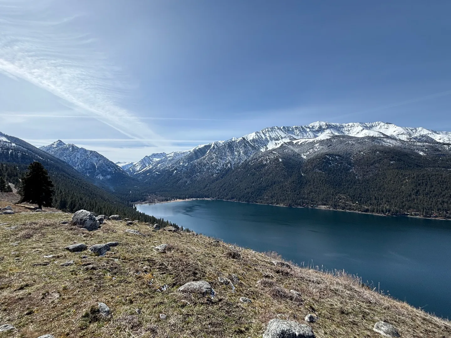 Elevated scenic overlook of a large deep blue lake bordered by evergreen-covered hills and snow-dusted mountains under a blue sky.