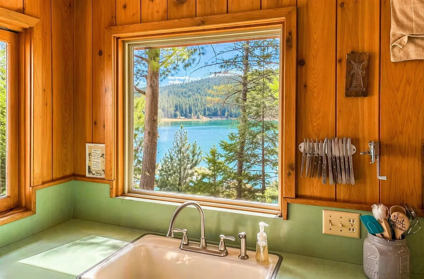 Kitchen sink beneath a window framing views of a blue lake and pine trees.