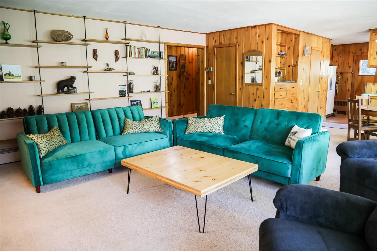 Living room featuring two teal tufted sofas arranged around a light wood coffee table with hairpin legs, backed by wood-paneled walls and floating shelves.