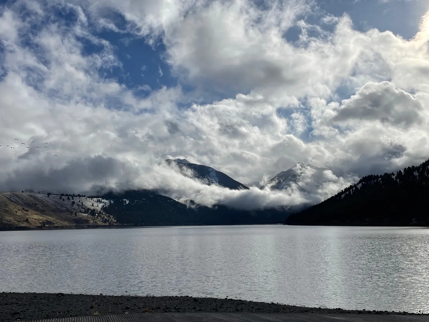 A wide view of a calm lake flanked by forested, snow-dusted mountains beneath a cloud-filled sky.