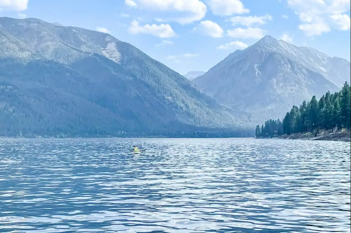 Wide view of a mountain lake with a small figure in a kayak or paddleboard, bordered by forested hills under a blue sky.