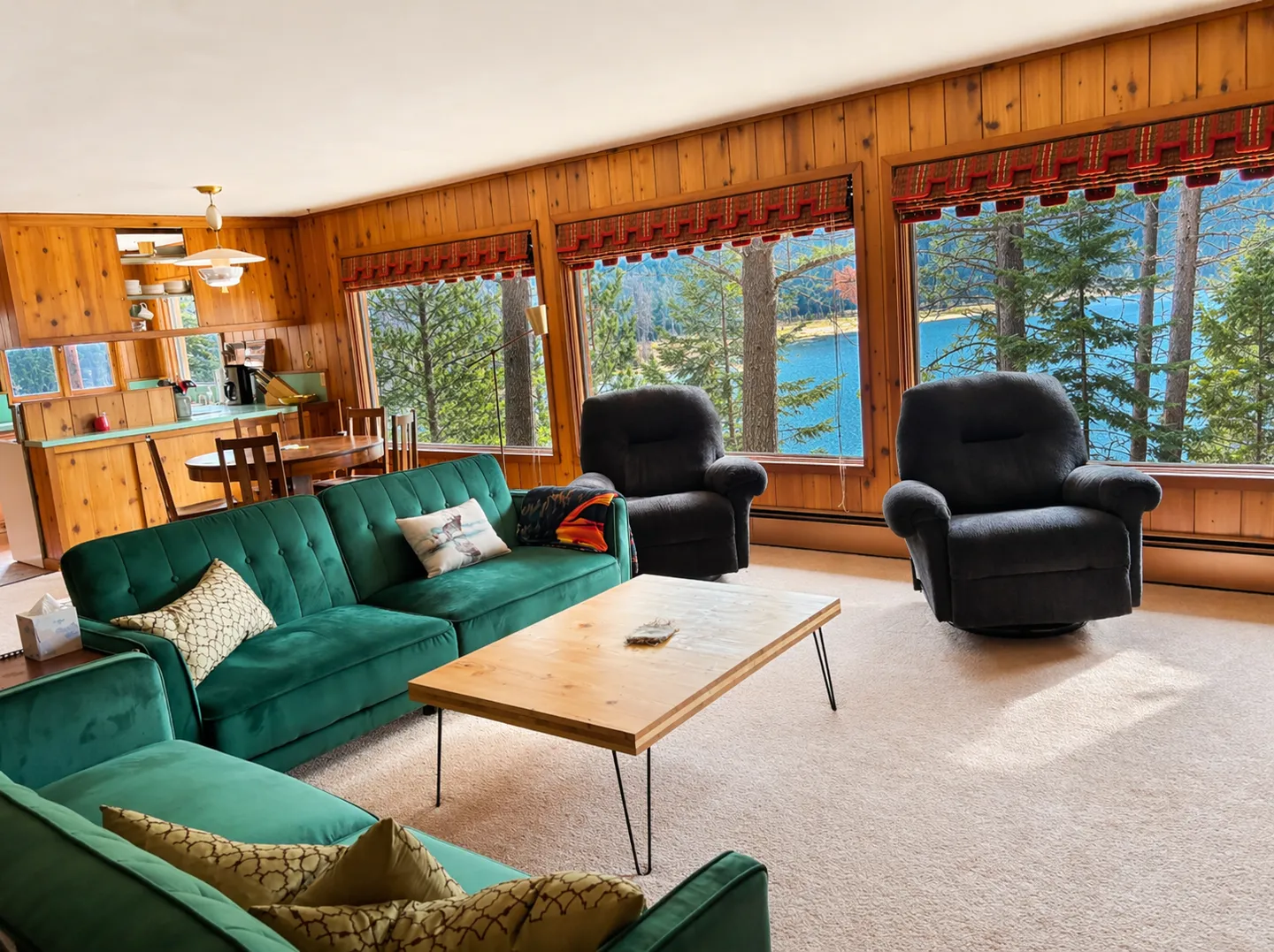 Living room with wood-paneled walls, green sofas, armchairs, and large windows looking out onto a lake through pine trees.