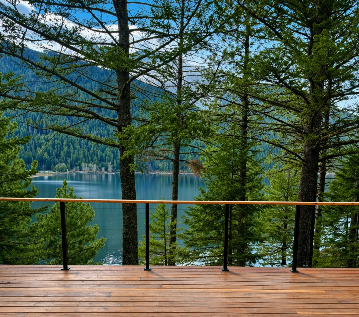 A wooden outdoor deck with a railing overlooking a calm lake surrounded by dense evergreen trees and mountains.