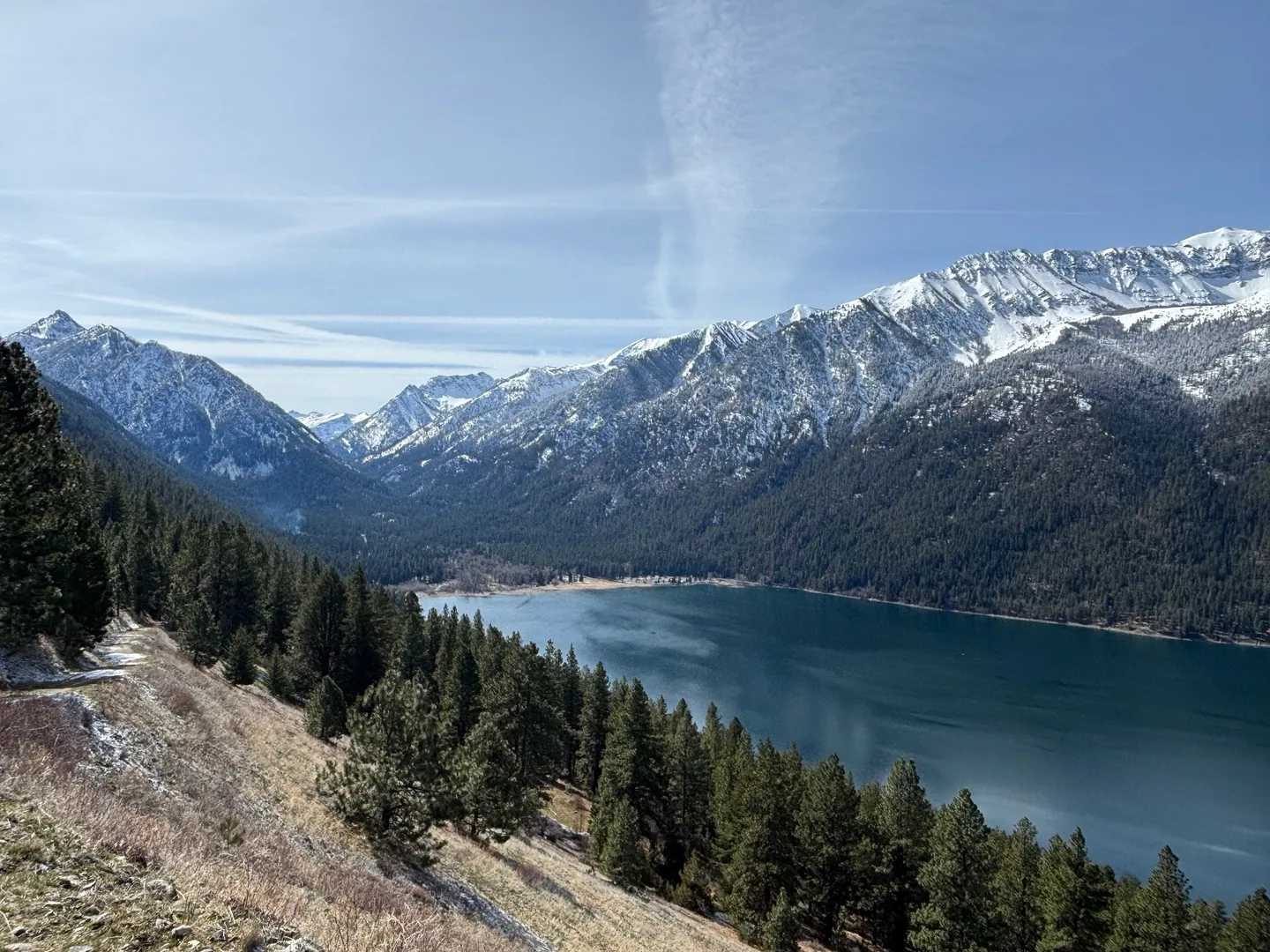 Elevated scenic view overlooking a large blue mountain lake surrounded by dense evergreen forests and snow-capped peaks under a blue sky.