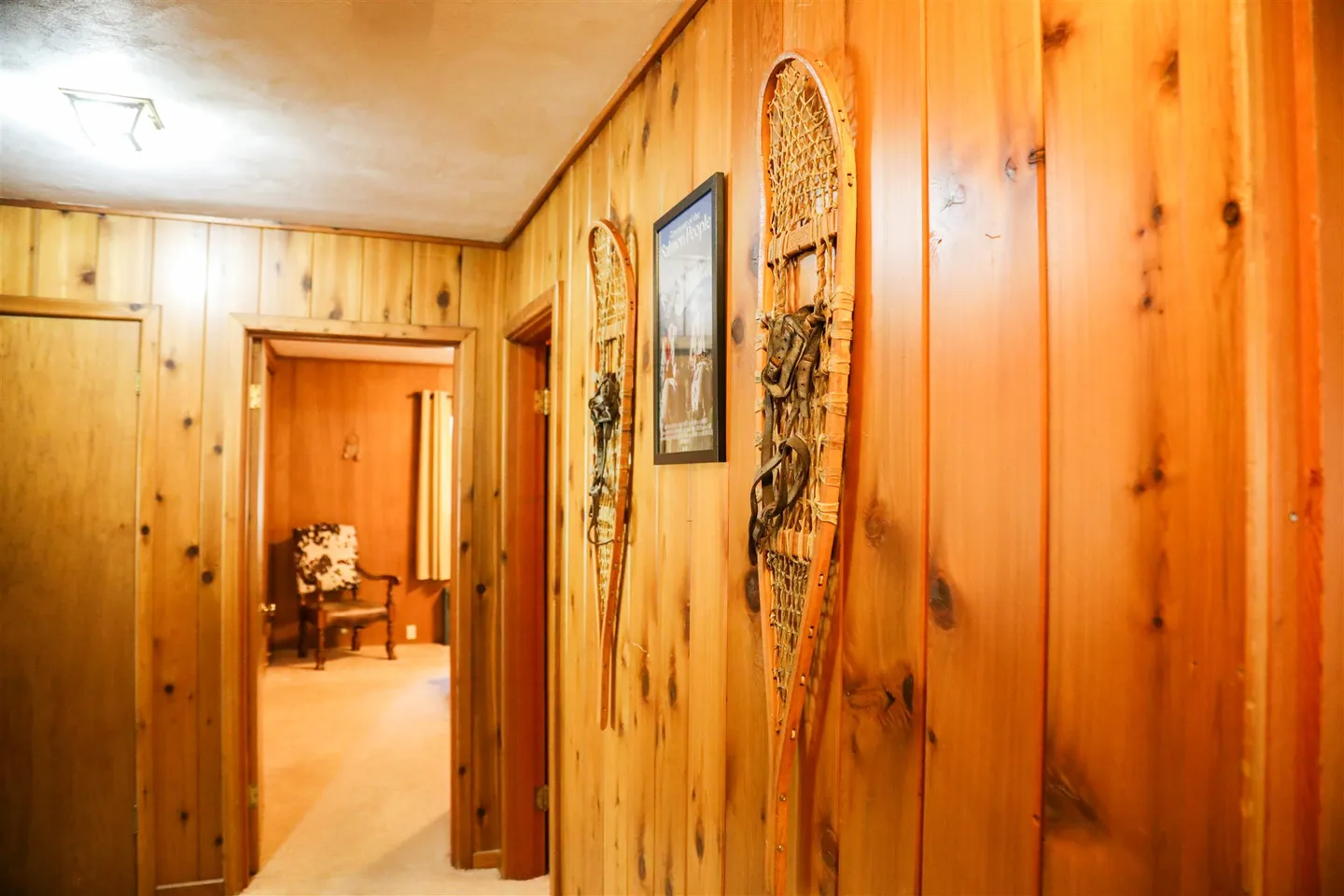Hallway featuring knotty pine wood-paneled walls decorated with vintage snowshoes and a framed picture, leading to an adjoining room.