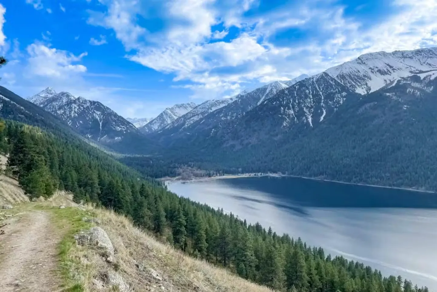 Elevated view from a dirt hiking trail looking over a lake surrounded by forested slopes and snow-dusted mountains under a partly cloudy blue sky.