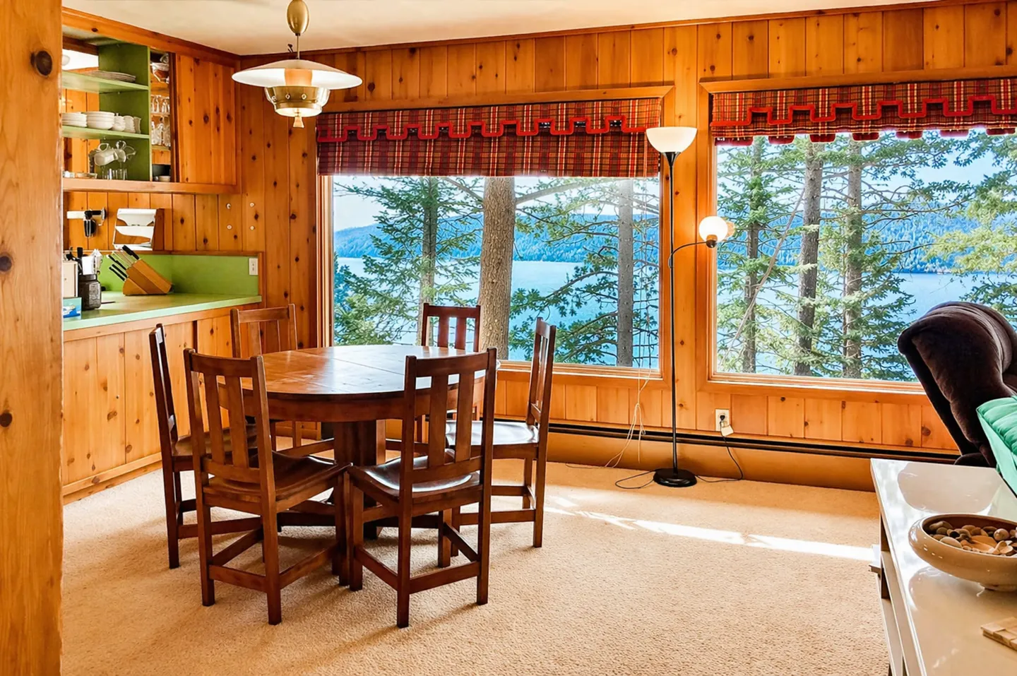 Dining area with wood-paneled walls, carpeted floor, round wooden table, and large picture windows looking out onto pine trees and a lake.
