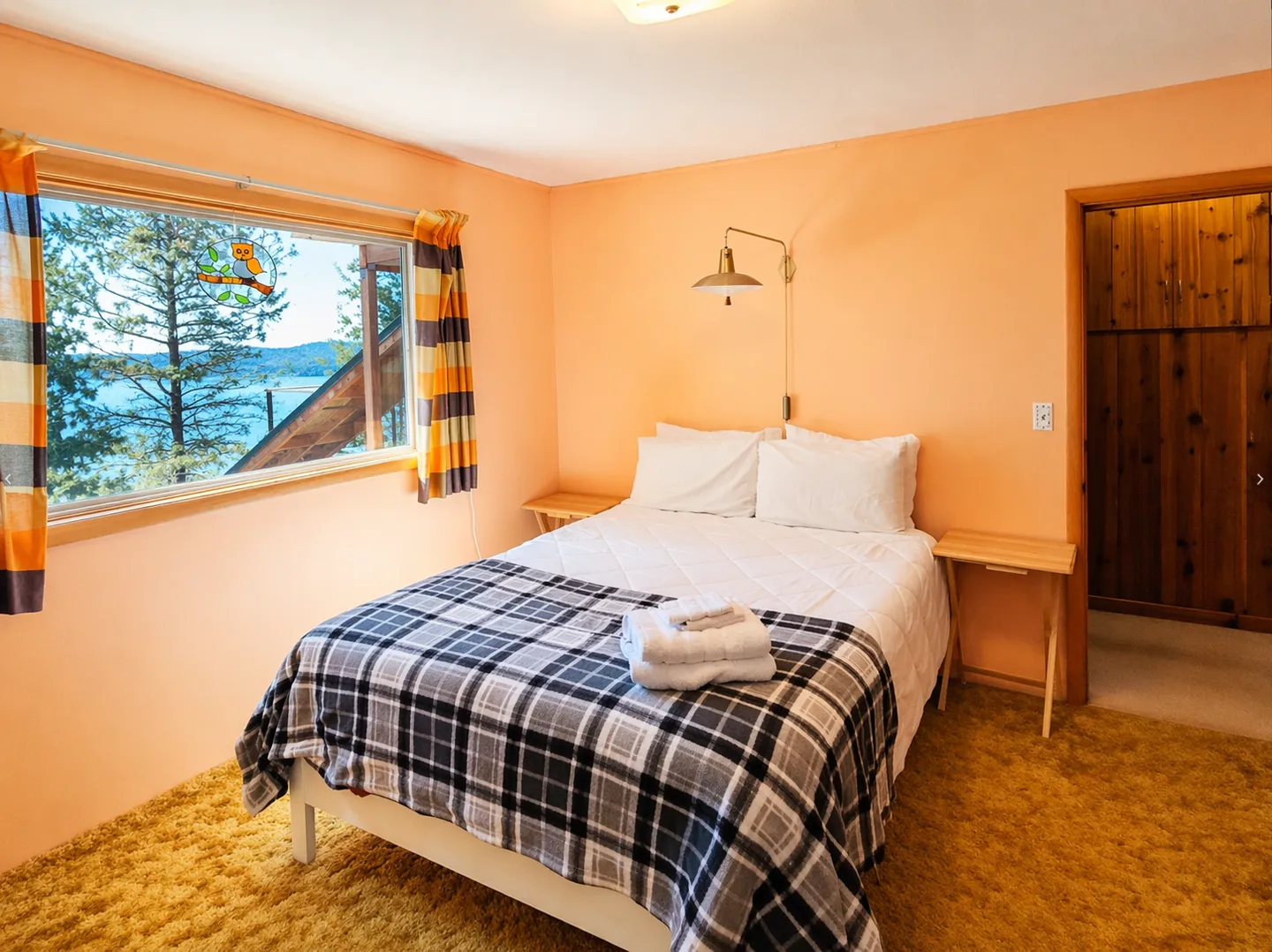 Bedroom with peach-colored walls, carpeted floor, a bed with a plaid throw, and a window overlooking a body of water and trees.
