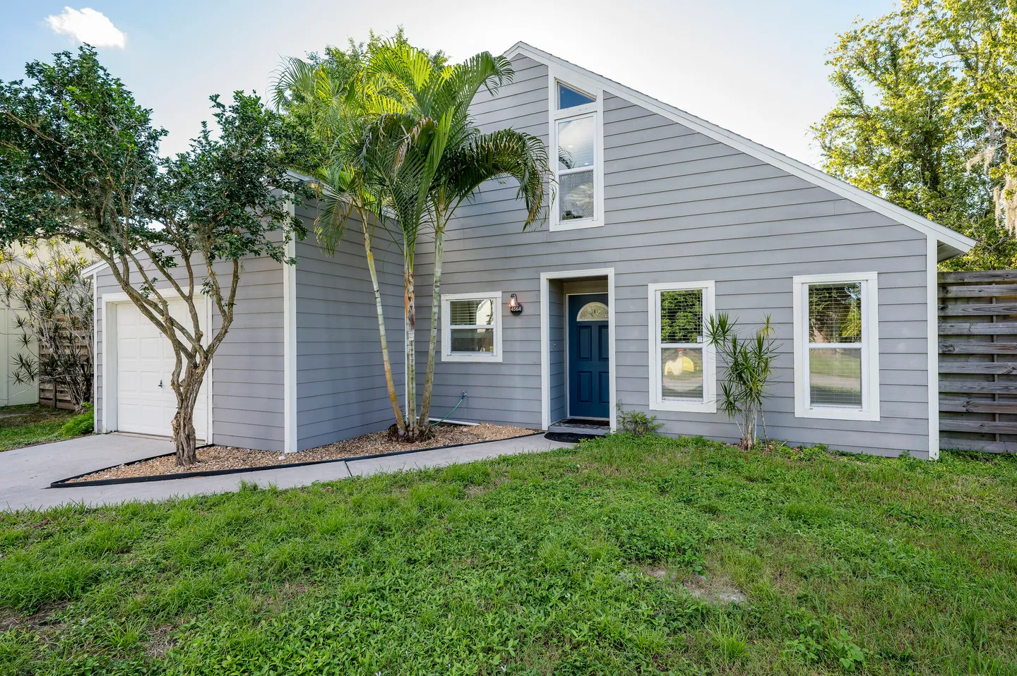 Front exterior of a gray siding home with a blue front door, garage, and front lawn with palm trees.