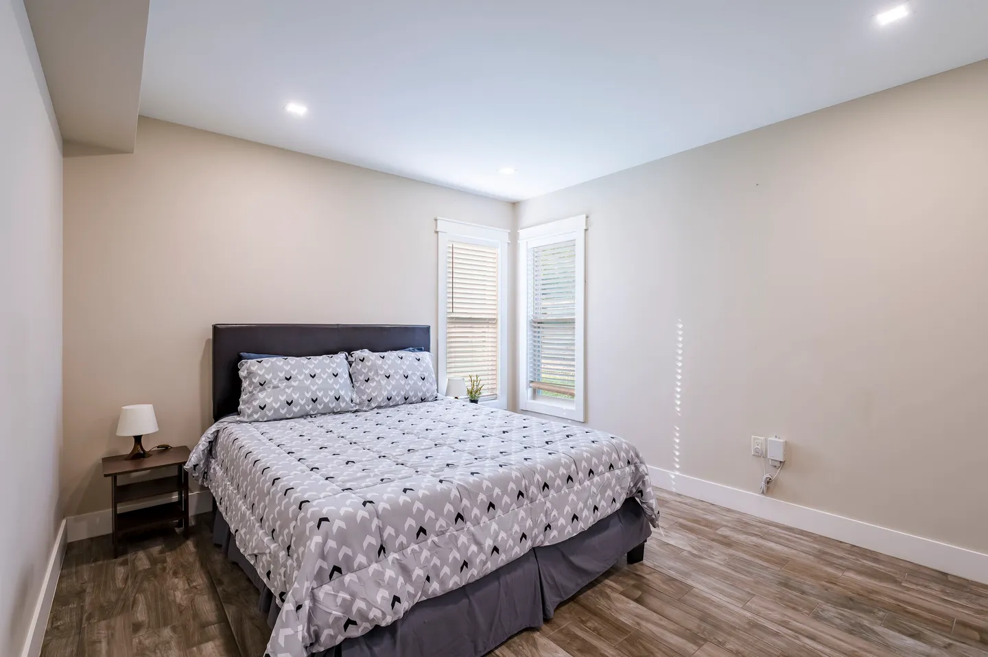 Bedroom with wood-look flooring, a dark headboard, patterned gray bedding, and two corner windows with blinds.