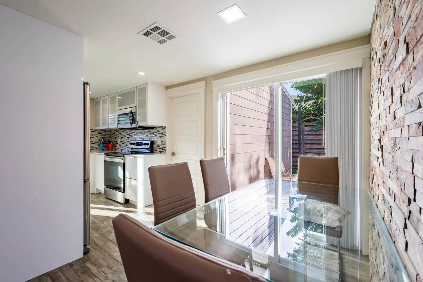 Dining area with a glass-top table, leather-style chairs, stone accent wall, and sliding glass door leading to a patio, with the kitchen in the background.