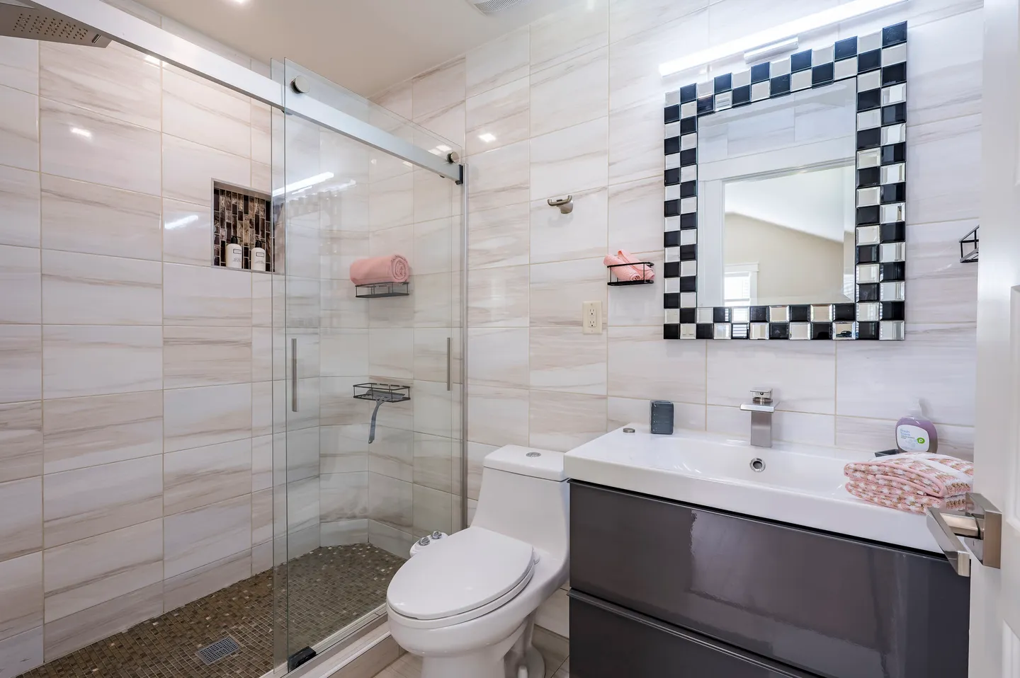 Bathroom with light-colored tile walls, a sliding glass door shower, a toilet, and a vanity with a checkerboard-framed mirror.