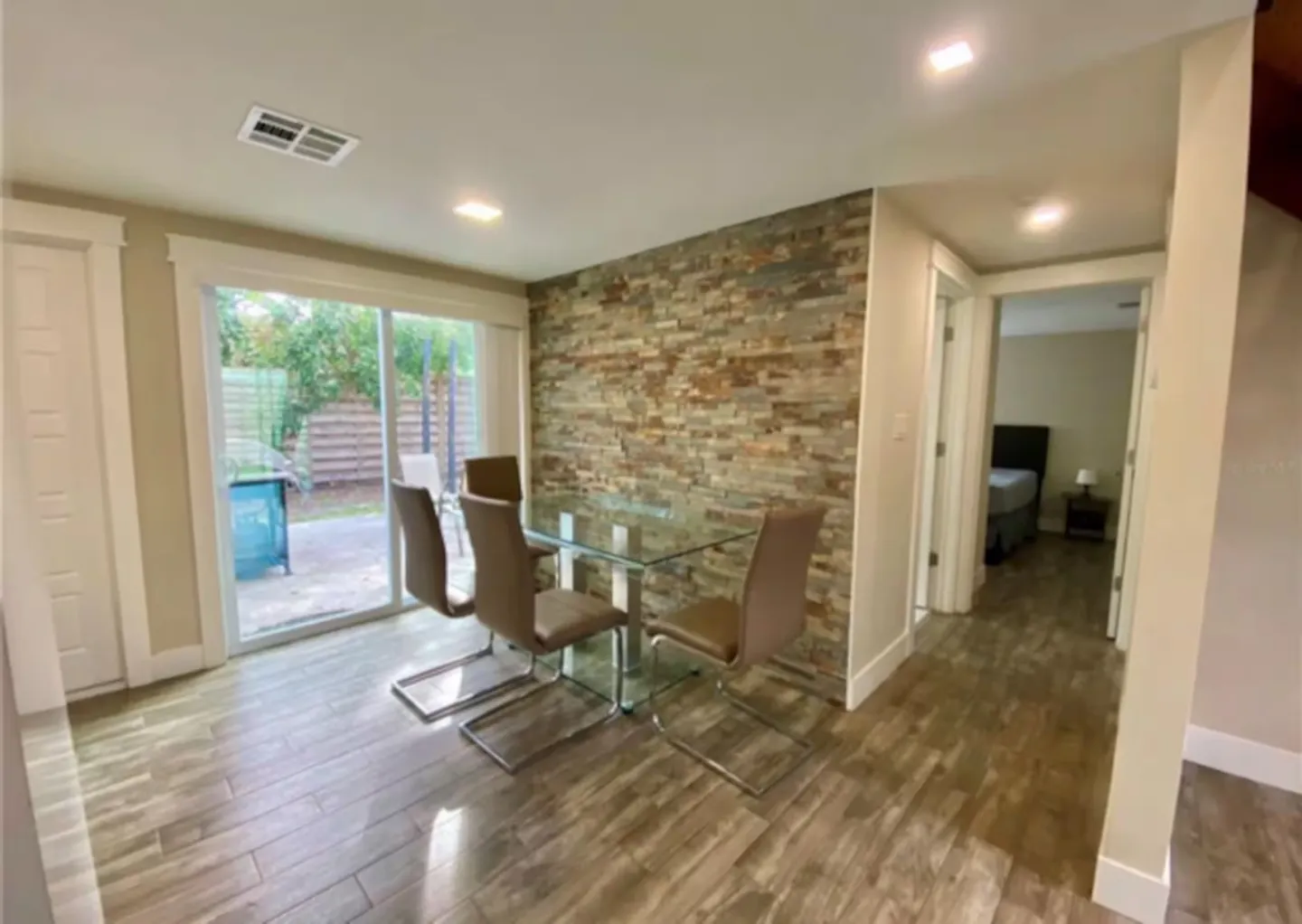 Dining area with a glass table and four chairs next to a stone accent wall and sliding glass door leading to a patio.