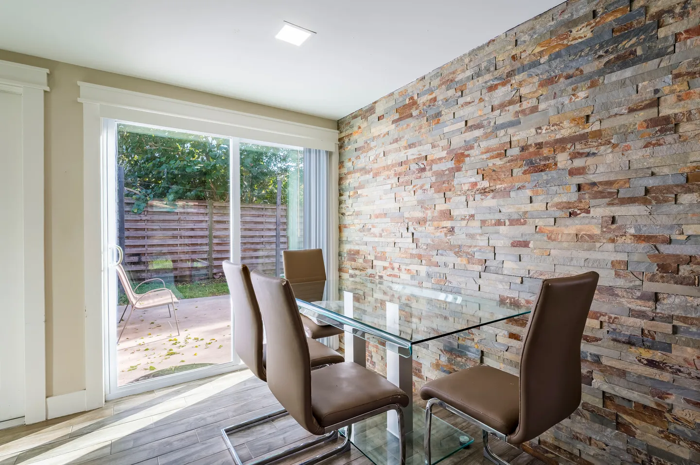 Dining area with a glass table, modern chairs, a stacked stone accent wall, and sliding glass doors leading outside.