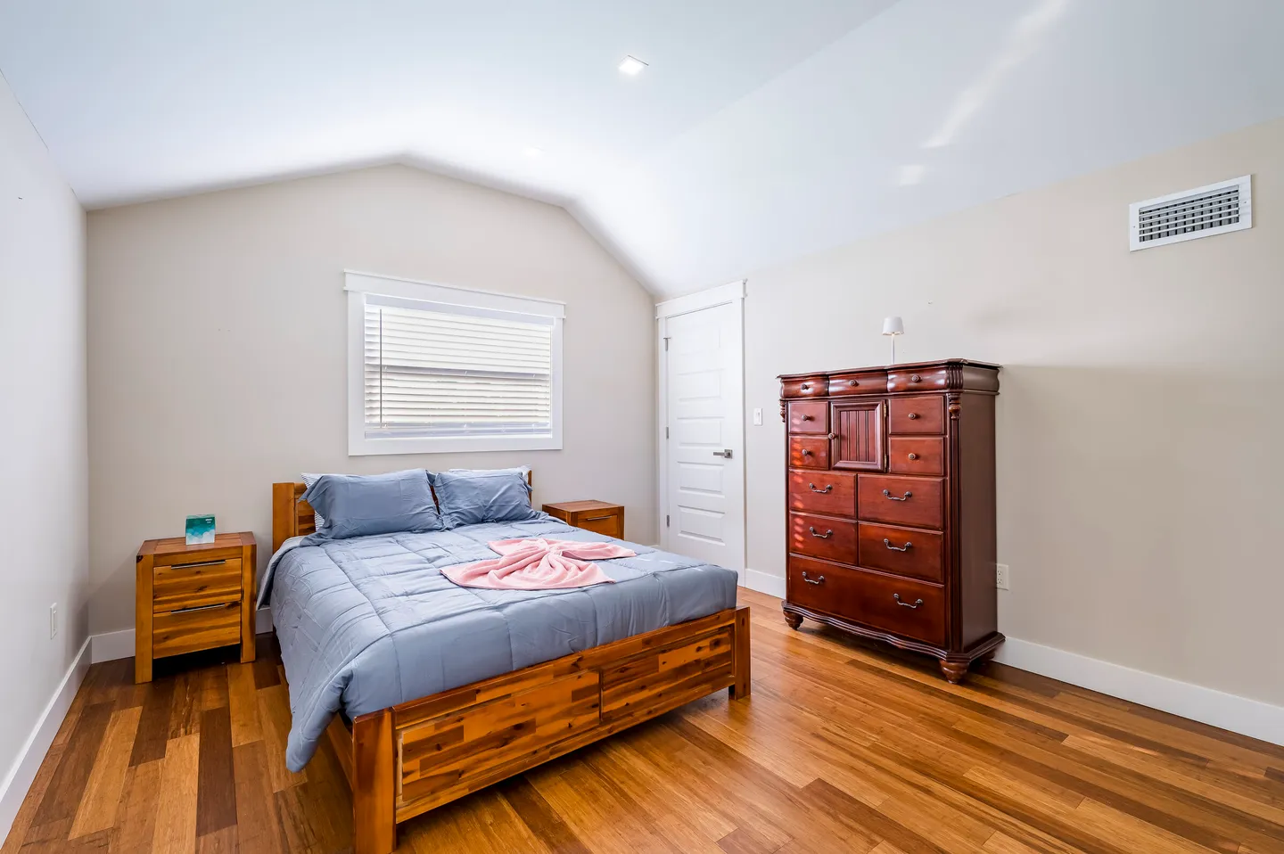 Bedroom with vaulted ceiling, wood-look flooring, a bed with a wooden frame, two nightstands, and a tall wooden dresser.