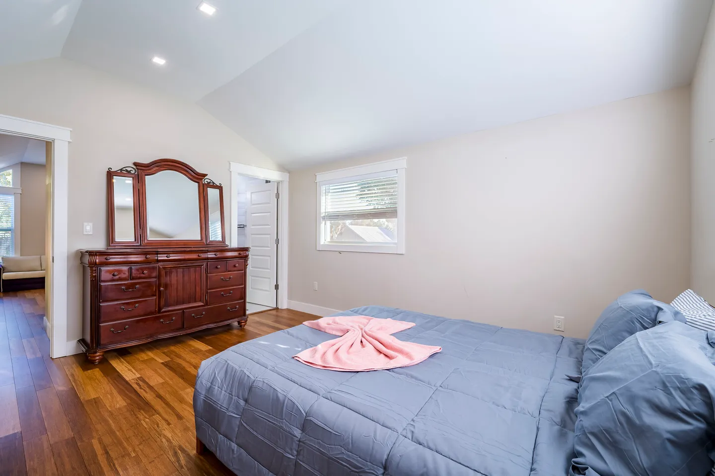 Bedroom with vaulted ceiling, wood-look flooring, a bed with blue bedding, and a large wooden dresser with attached mirrors.