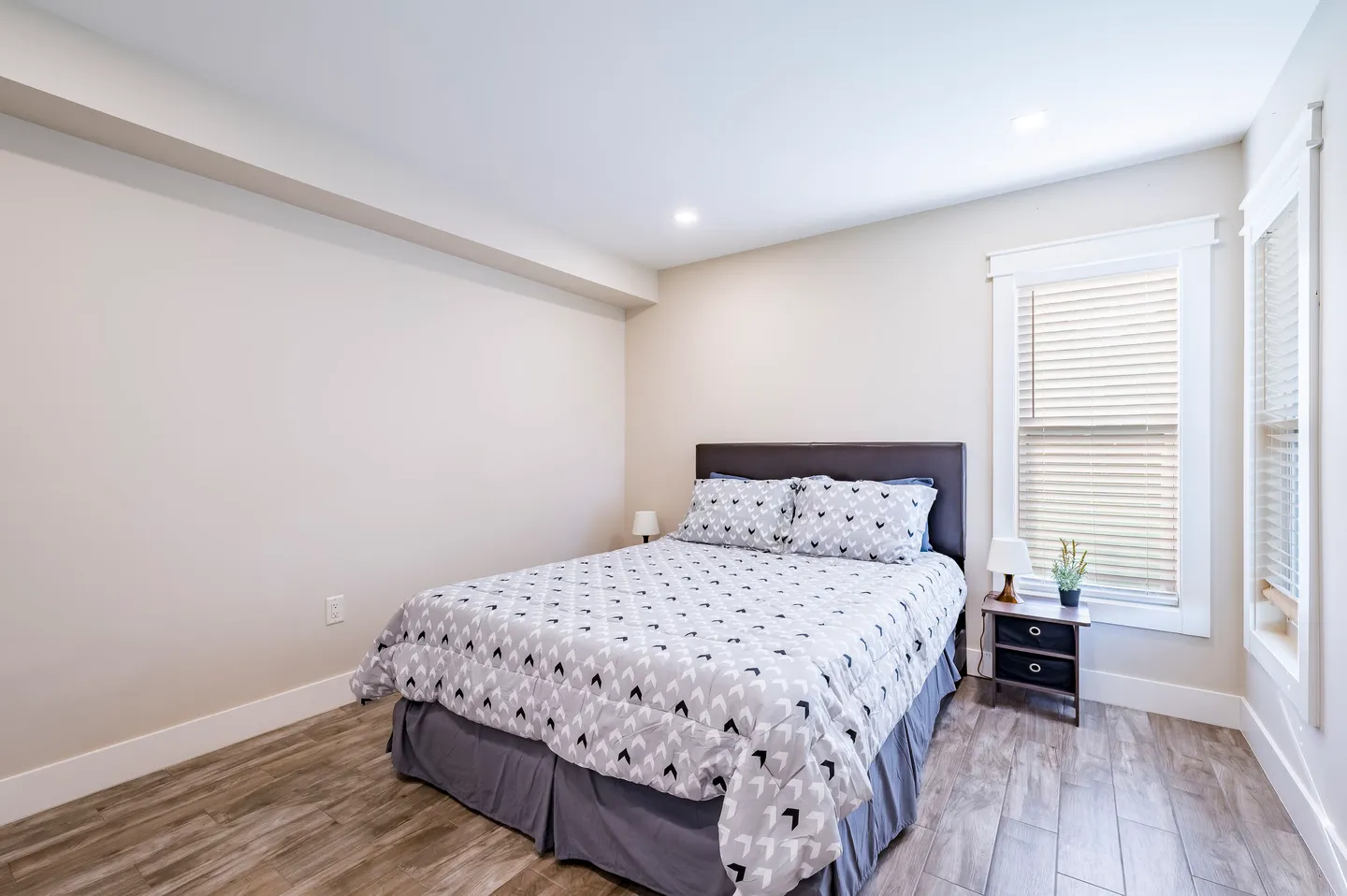 Bedroom with wood-look flooring, a made bed with patterned gray bedding, and a window with blinds.