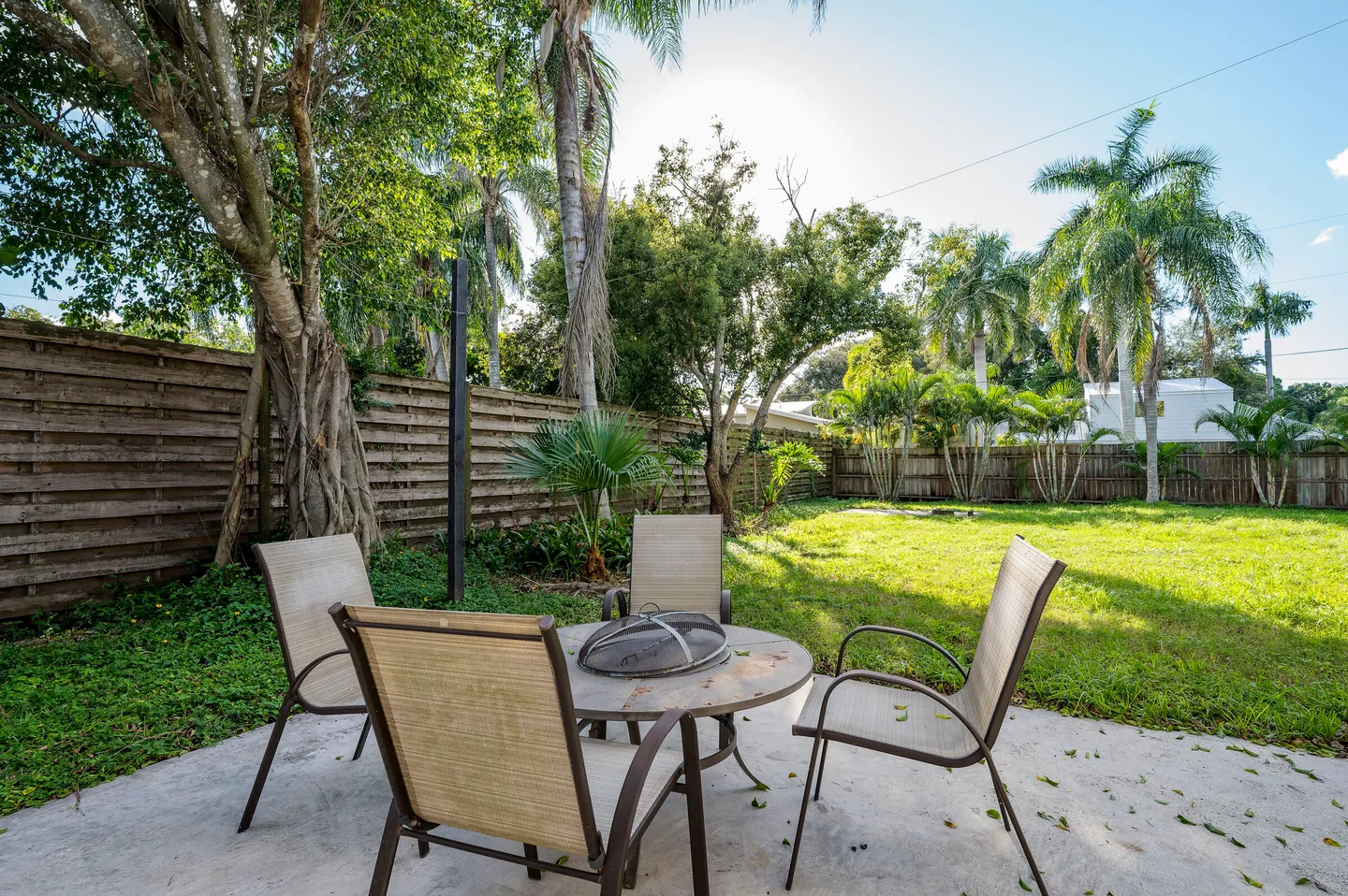 A concrete patio with outdoor chairs and a fire pit table in a grassy backyard enclosed by a wooden fence with palm trees.