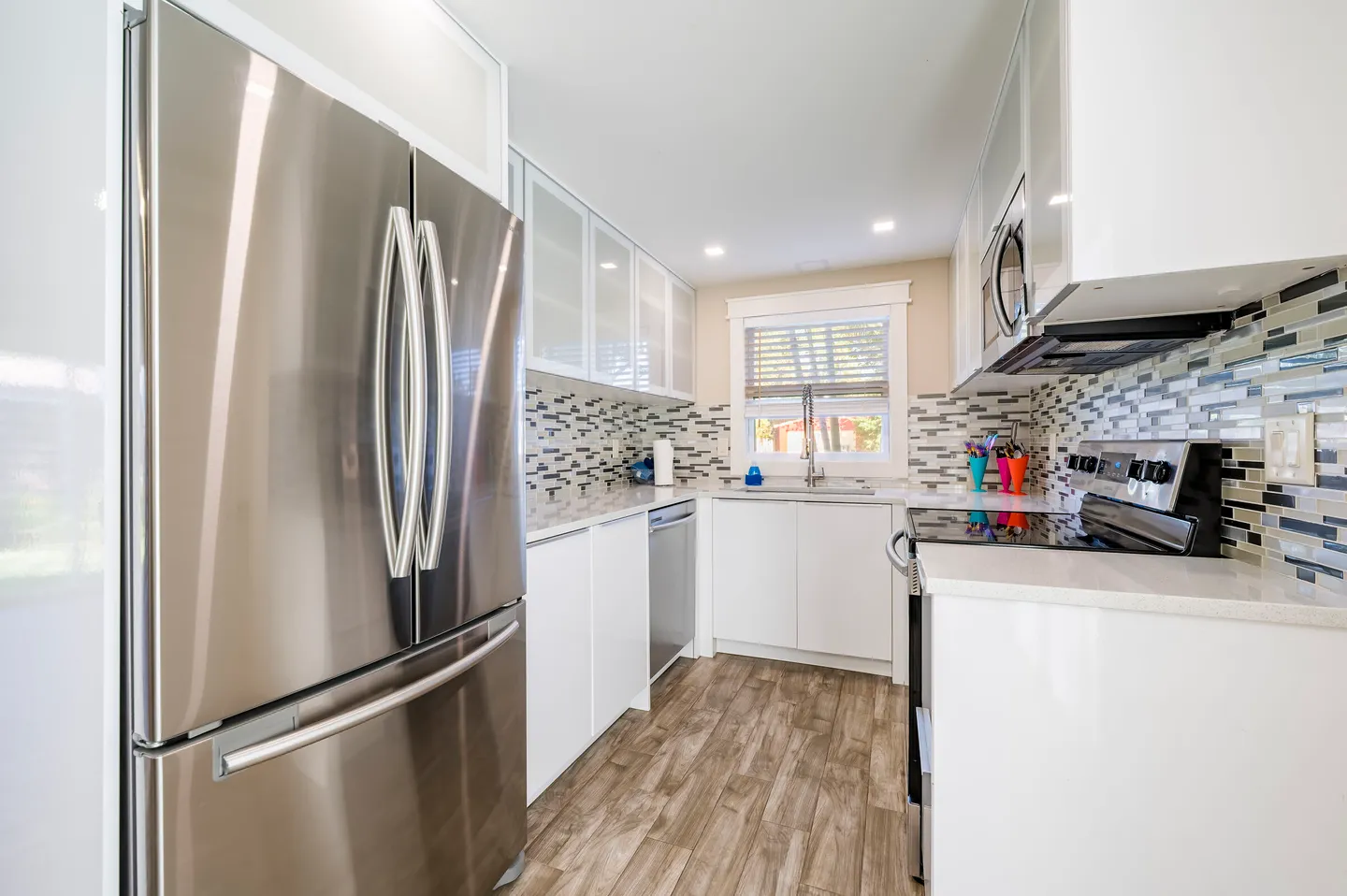 Bright kitchen featuring white cabinets, stainless steel appliances, a multi-colored tile backsplash, and light wood-look flooring.