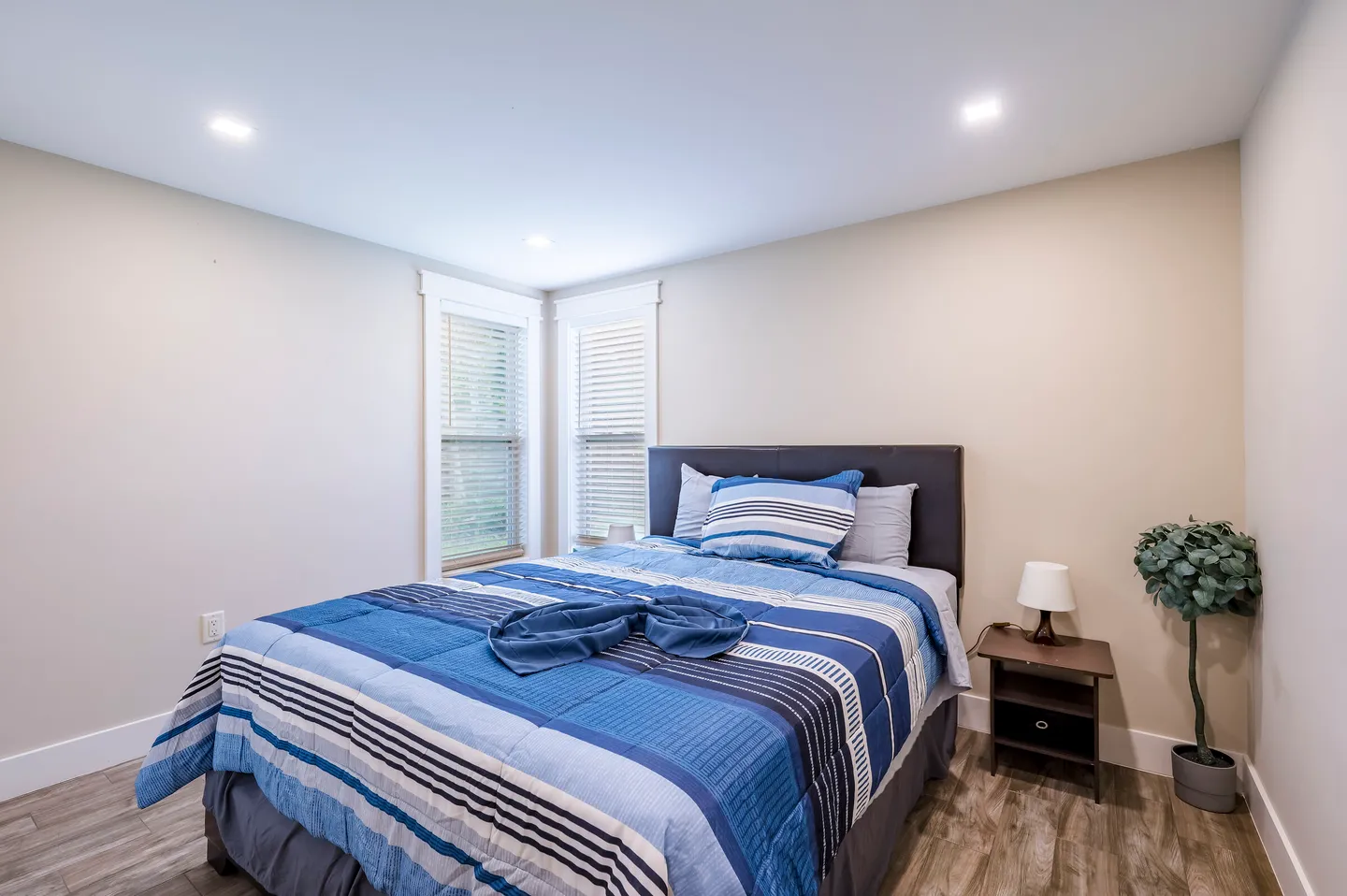 Bedroom with a blue striped comforter on the bed, wood-style flooring, a dark nightstand with a small lamp, and two corner windows with blinds.