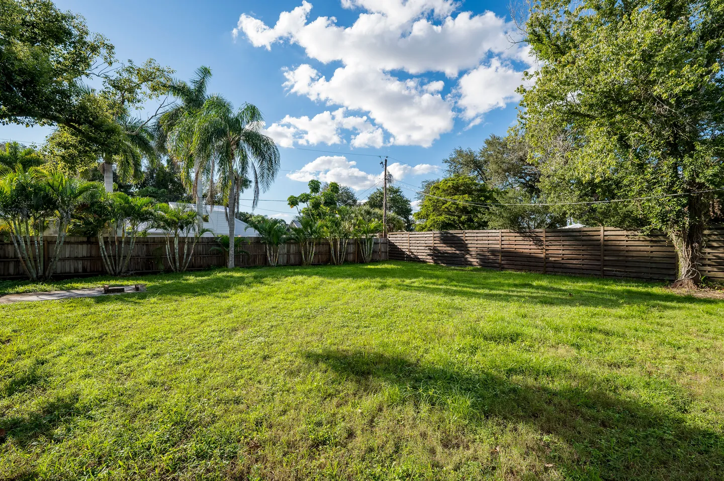 A large, fenced-in grassy backyard with palm trees under a bright blue sky with scattered clouds.