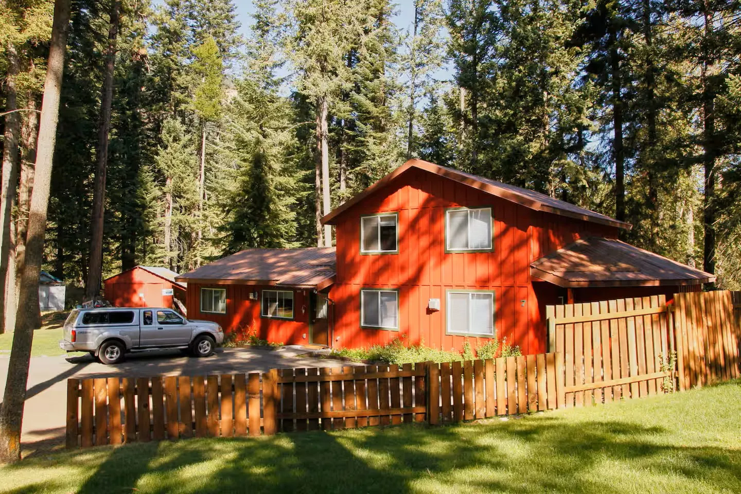 Exterior view of a red two-story home surrounded by tall evergreen trees with a wooden fence and parked truck in front.