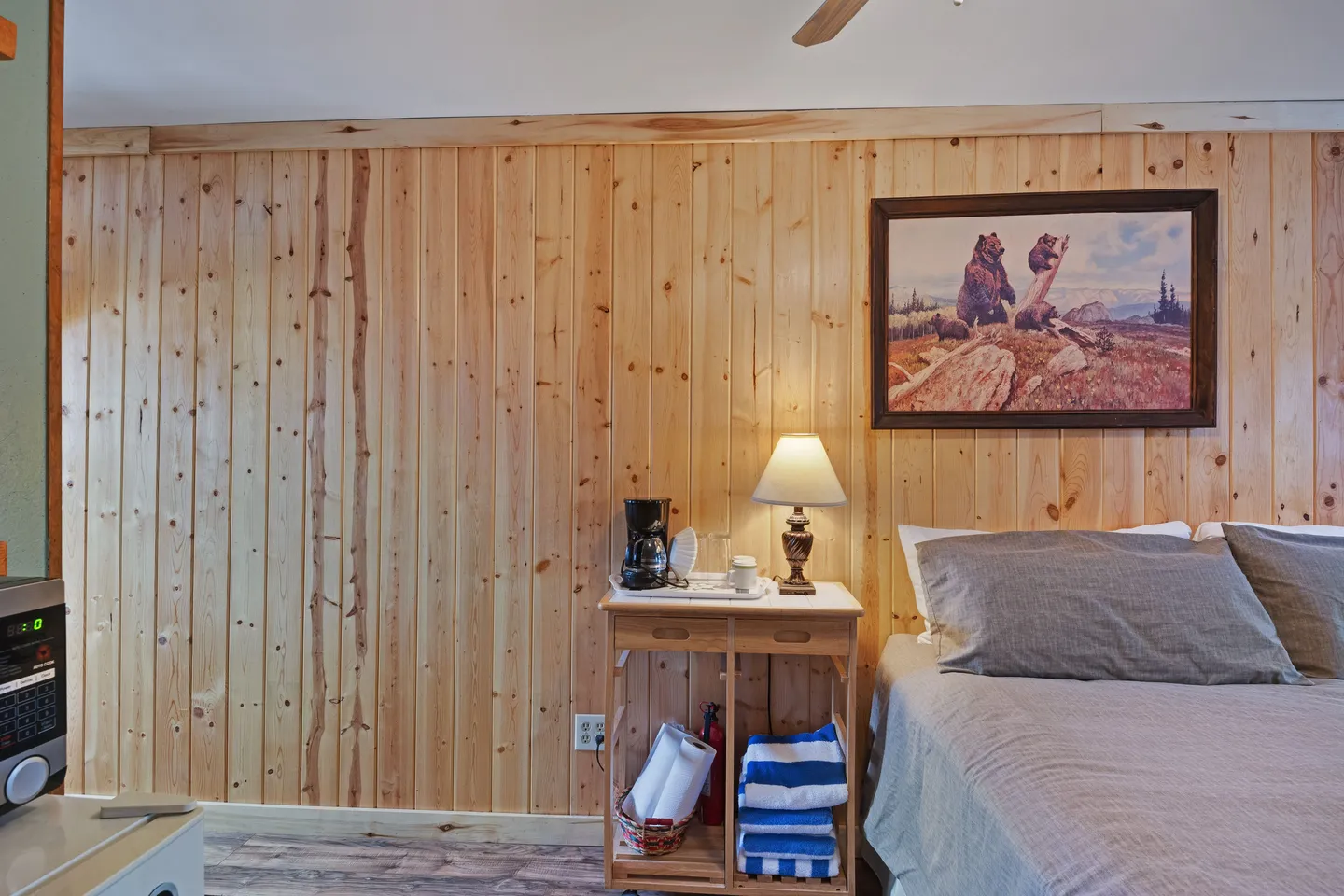 Bedroom with vertical pine-paneled walls, a bed, a nightstand holding a lamp and coffee maker, and bear artwork on the wall.