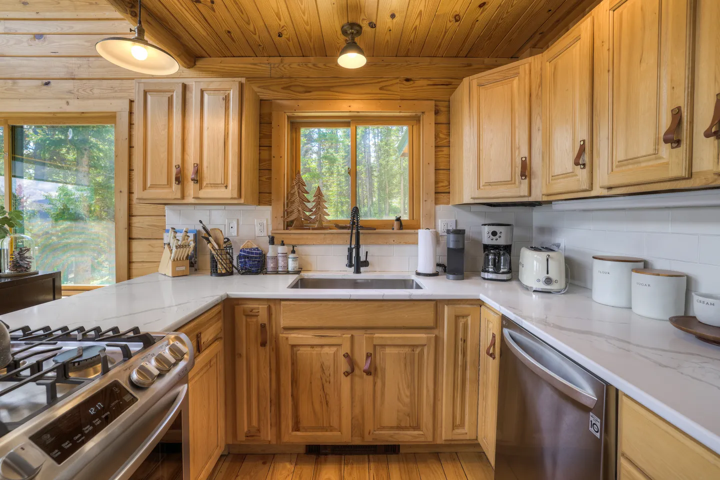 Kitchen with light wood cabinets, white countertops, stainless steel appliances, and a window above the sink looking out to trees.