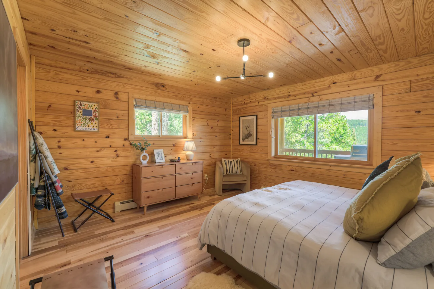 Bedroom with knotty wood paneled walls and ceiling, a bed with pillows, dresser, armchair, and windows overlooking greenery.