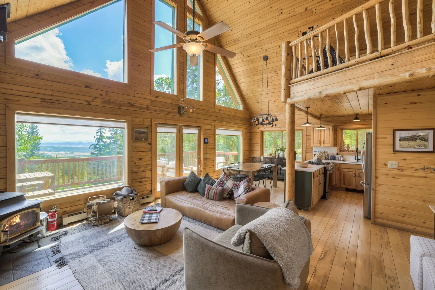 Open-concept log cabin living room with soaring vaulted ceilings, large windows overlooking outdoor scenery, a wood-burning stove, and adjacent kitchen and loft.