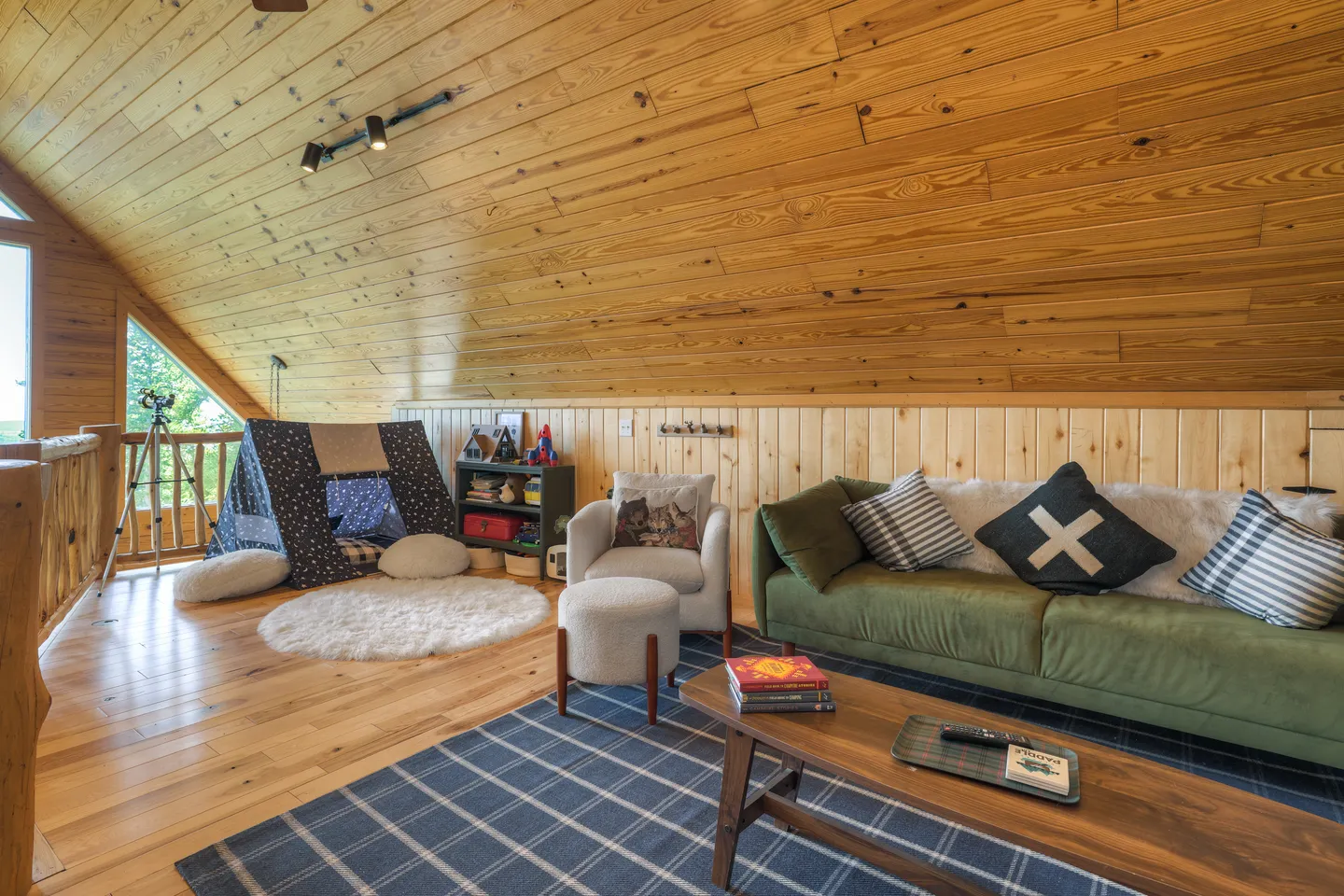 Cozy loft area with sloped wood-paneled ceilings, a green couch, an armchair, a play tent, and a telescope near the window.