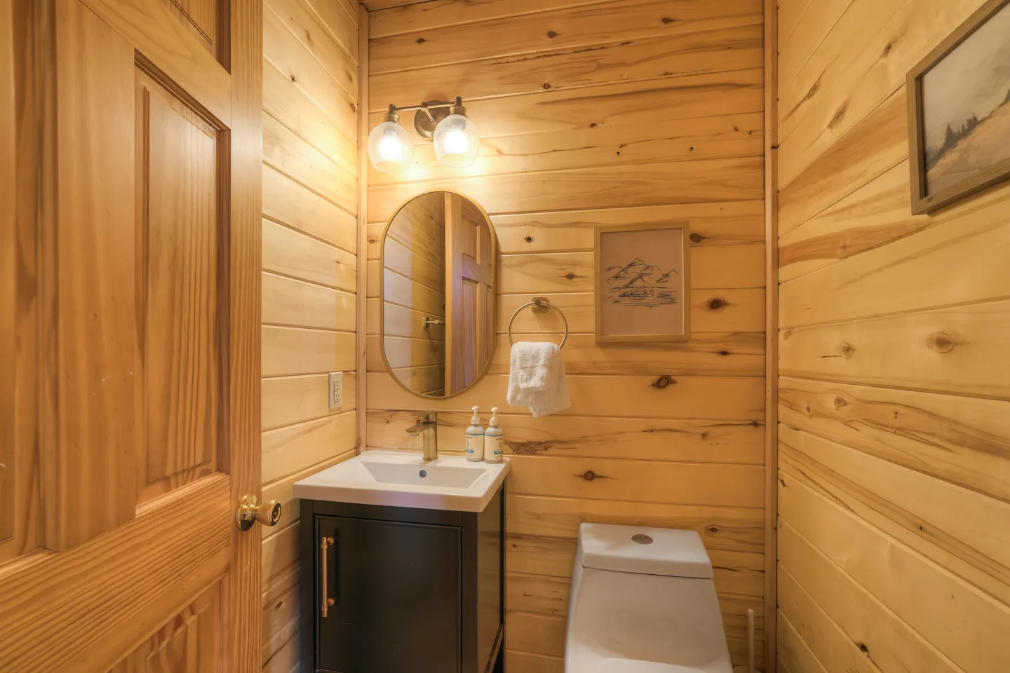 Powder room with wood-paneled walls, a dark vanity, an oval mirror, a modern toilet, and twin wall sconces.