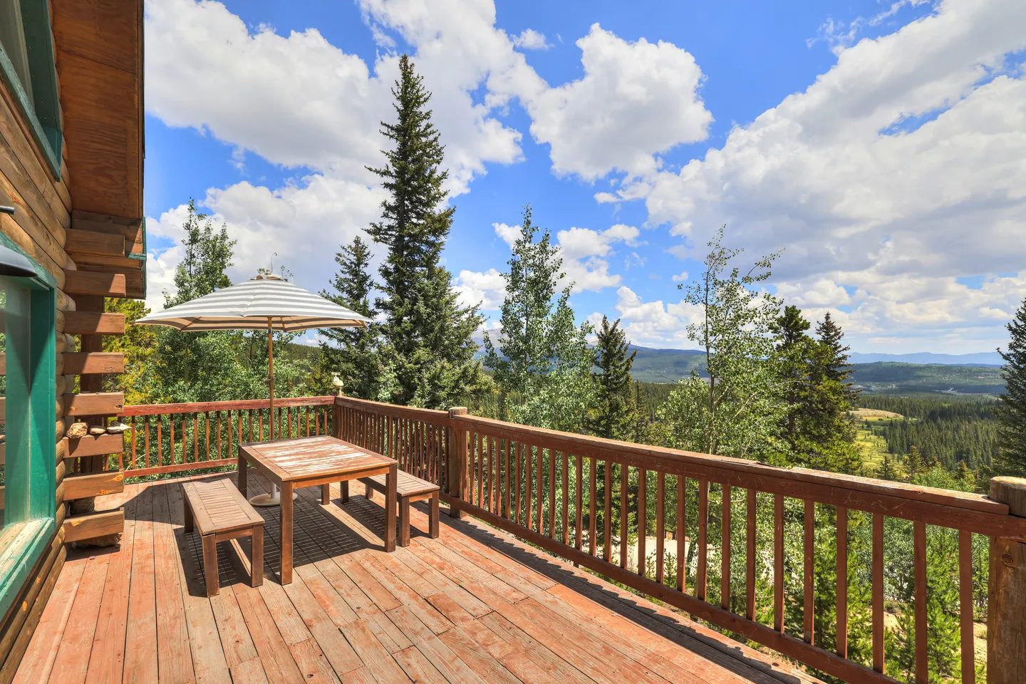 Outdoor wooden deck with a picnic table and umbrella overlooking evergreen trees and distant mountains under a blue sky with clouds.