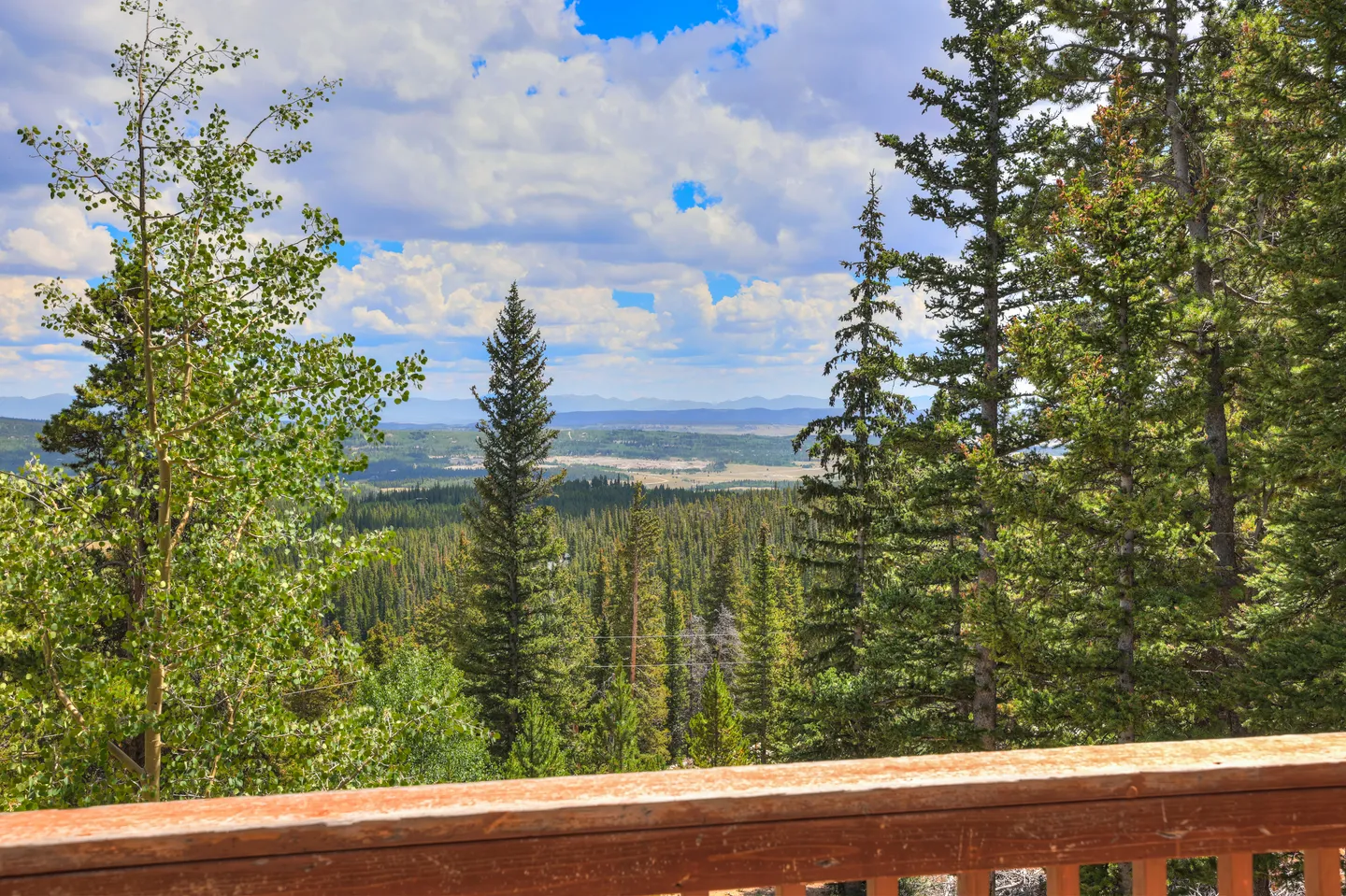 Elevated view from a wooden deck railing looking over tall evergreen trees toward a valley and distant mountains under a partly cloudy sky.