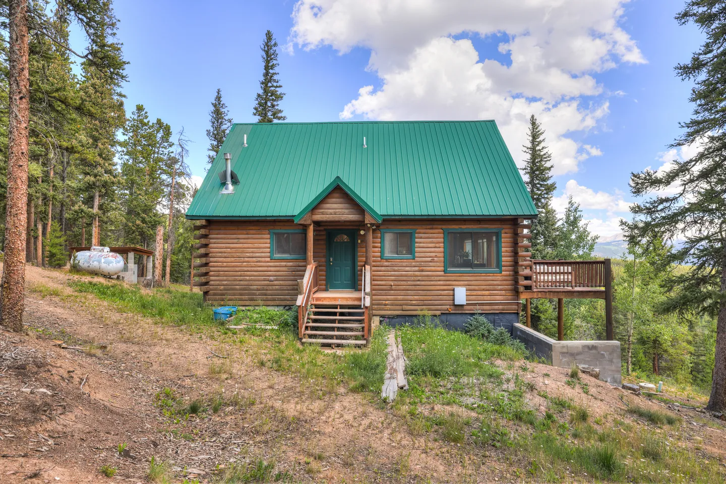 Exterior view of a log cabin with a bright green metal roof, front steps, side deck, and surrounding pine trees.