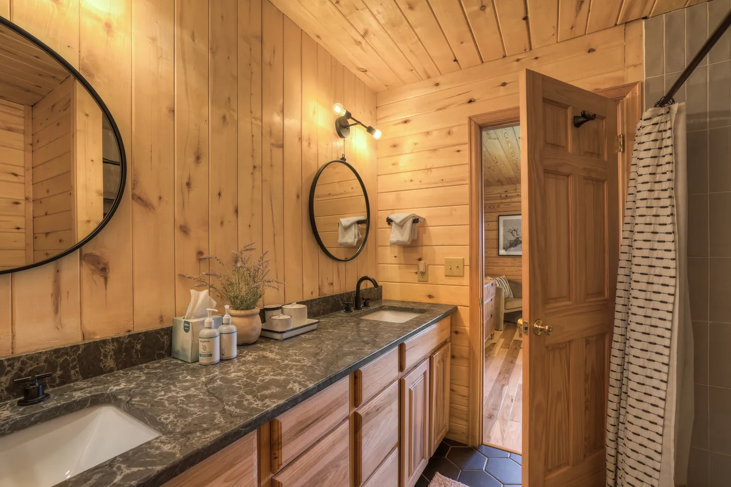 Bathroom with wood-paneled walls, a double-sink dark countertop vanity, round mirrors, and an open doorway showing a bedroom.