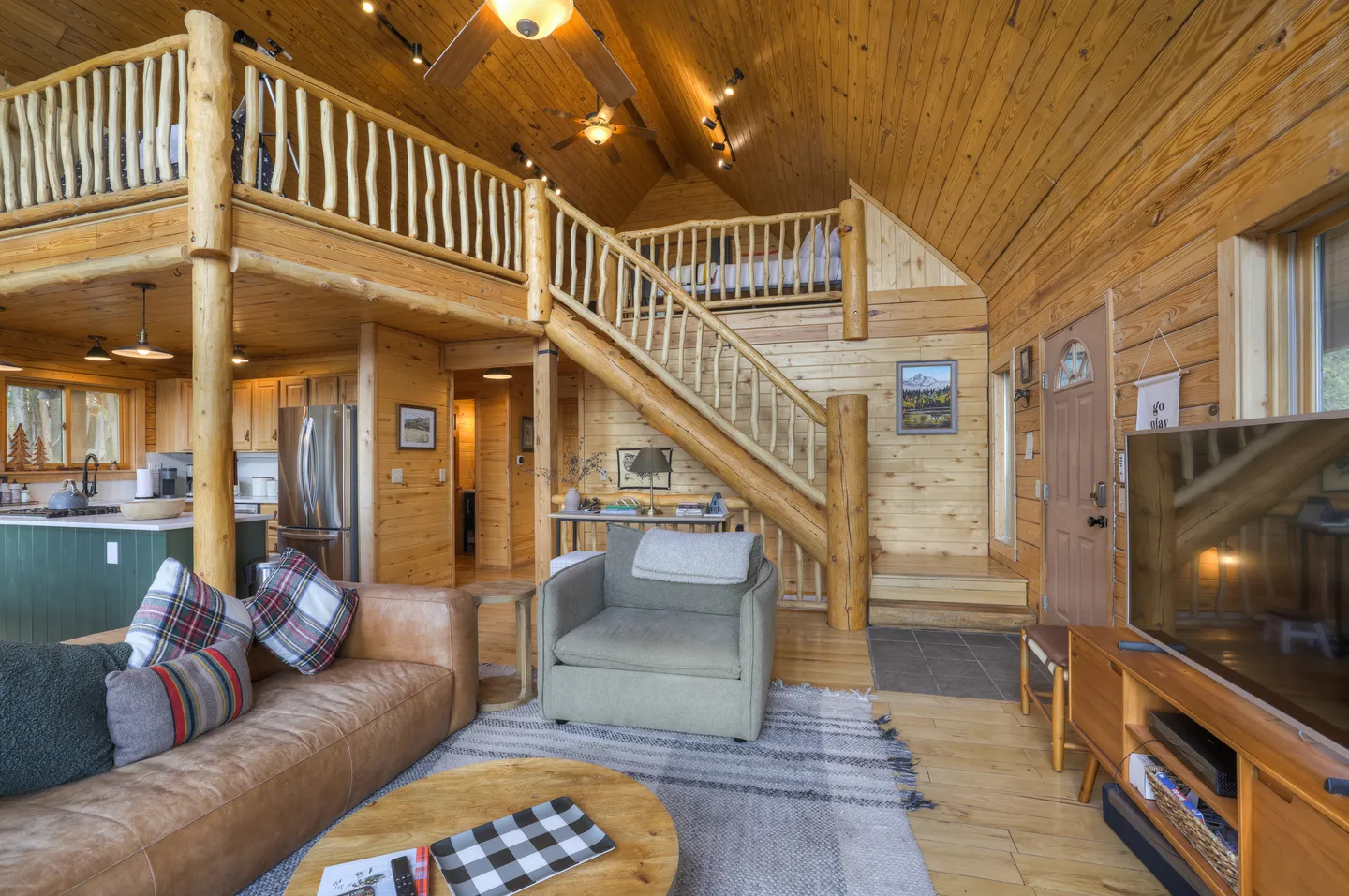 Open-concept cabin living room with log railings, loft area, wood-paneled walls and ceiling, and a view into the kitchen.