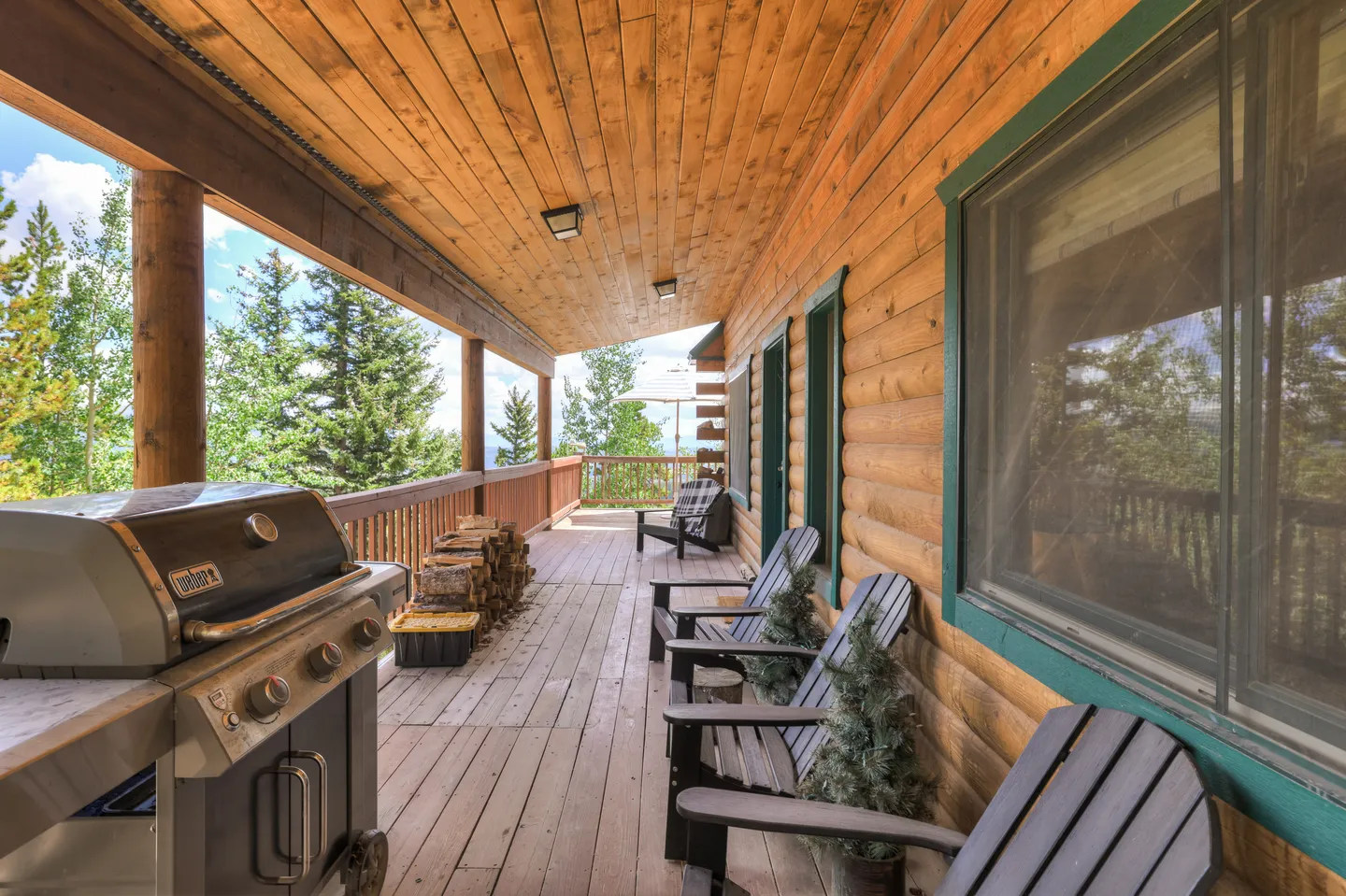 A covered wooden deck on a log home featuring a gas grill, stacked firewood, and Adirondack chairs overlooking surrounding trees.