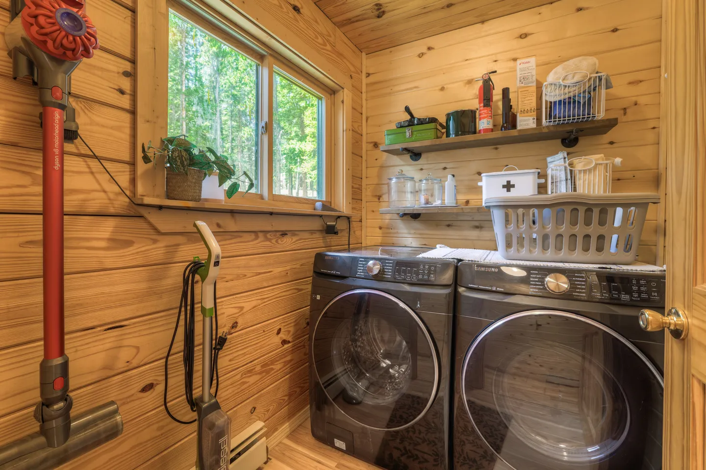 A laundry room with wood paneled walls, a front-loading washer and dryer, open shelving, and a window.