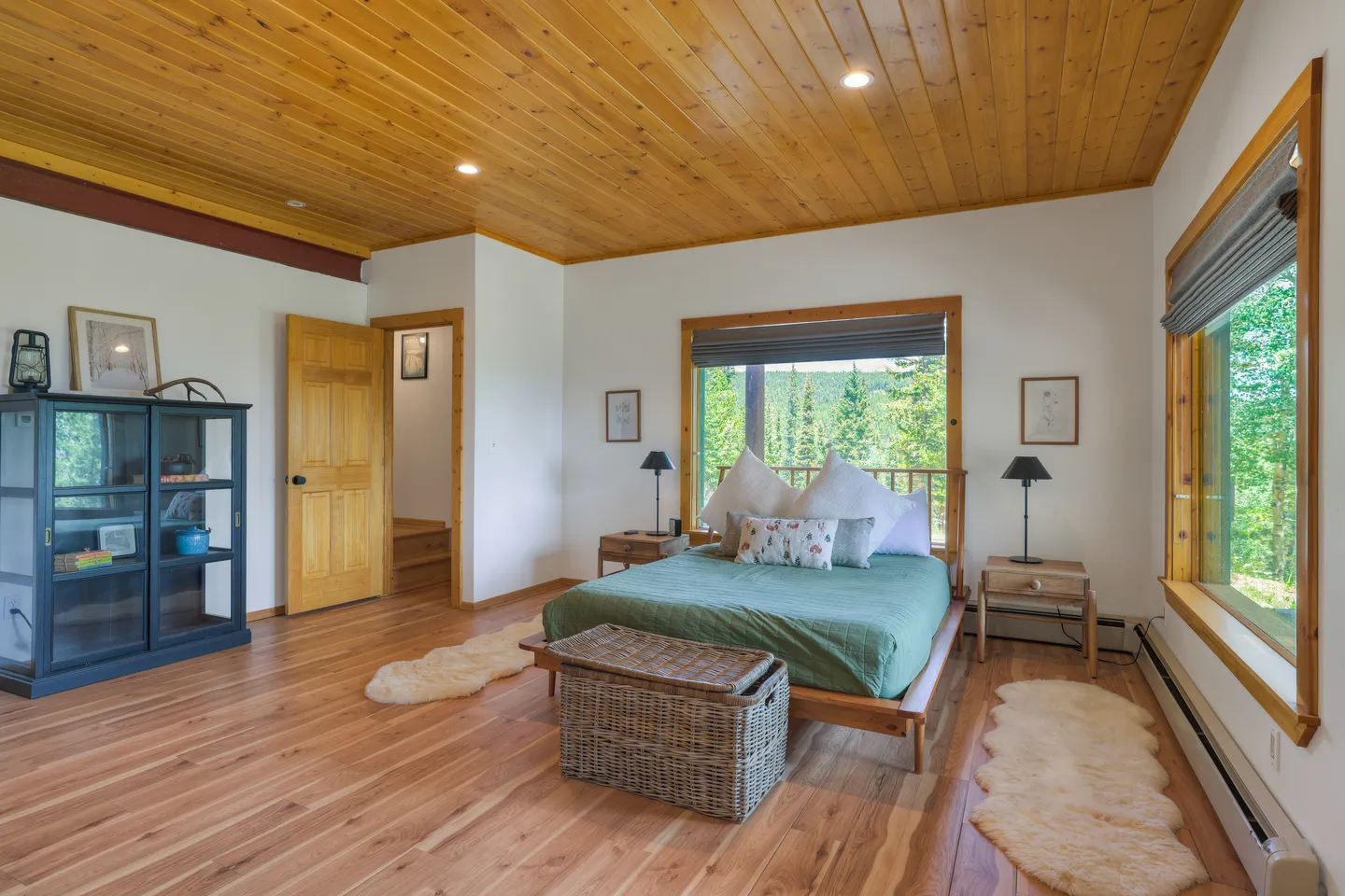 Bedroom with wooden ceiling, wood floor, made bed with green duvet, and large windows looking out at pine trees.