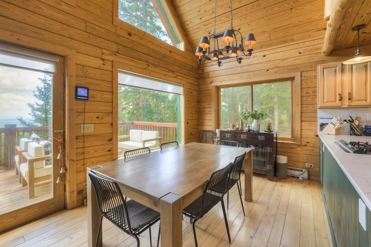 Dining area with a wooden table, black chairs, wood-paneled walls, chandelier, and windows looking out to an outdoor deck.