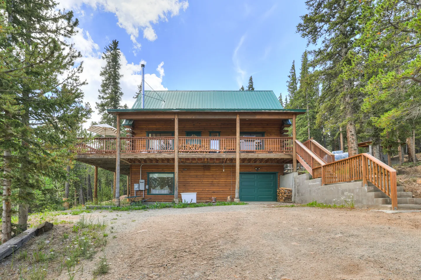 Exterior of a two-story log home with a green metal roof, wrap-around upper deck, single garage door, and gravel driveway surrounded by pine trees.