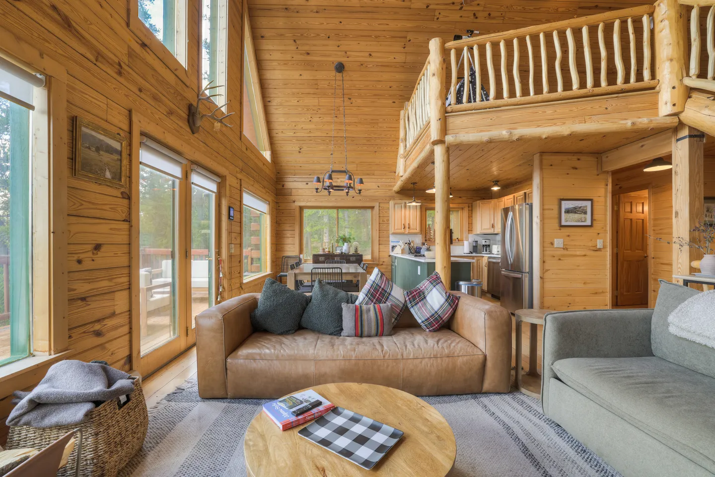Open-concept log cabin interior with high vaulted wood ceilings, a living room with sofas, an overlooking loft, and the kitchen and dining area in the background.