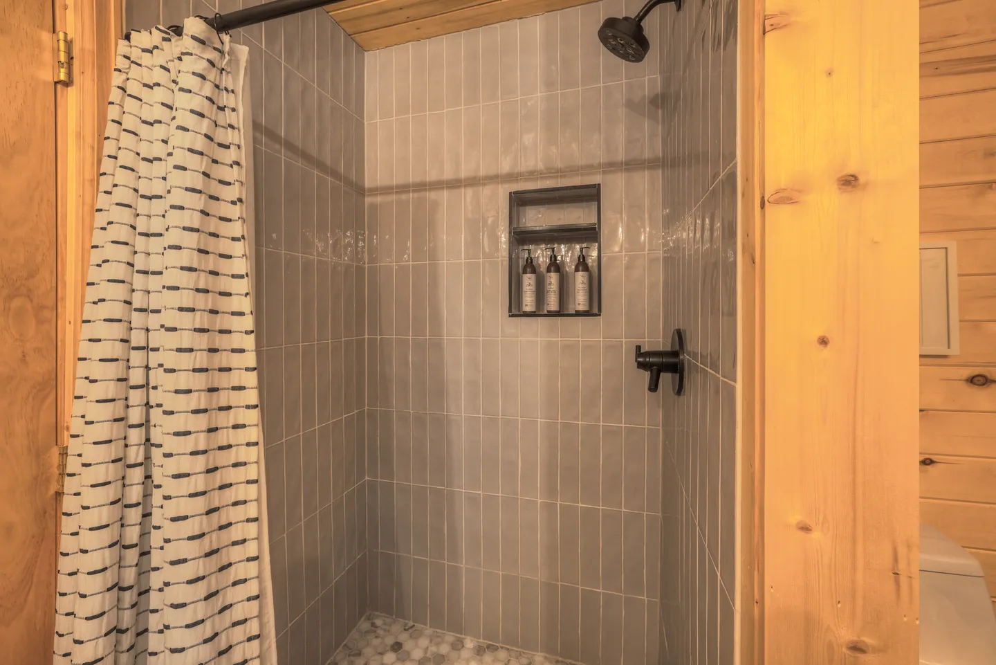 Tiled walk-in shower with dark hardware, a built-in wall niche, and a patterned shower curtain.