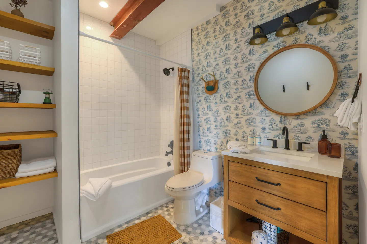 Bathroom with a wood vanity, round mirror, toilet, white-tiled shower-tub combo, and blue patterned wallpaper.
