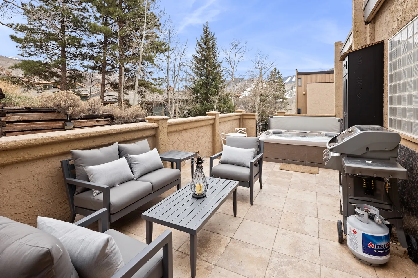 Outdoor patio with tiled floor, modern cushioned lounge seating, a coffee table, a hot tub, and a barbecue grill with views of trees and hills.