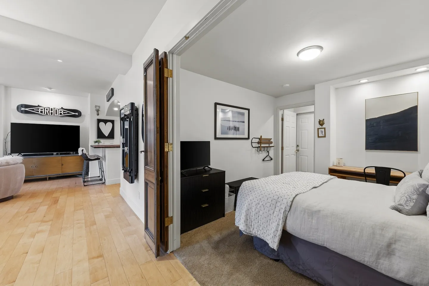 View through double doors into a bedroom with a bed and dresser, adjacent to a living room with light wood floors.