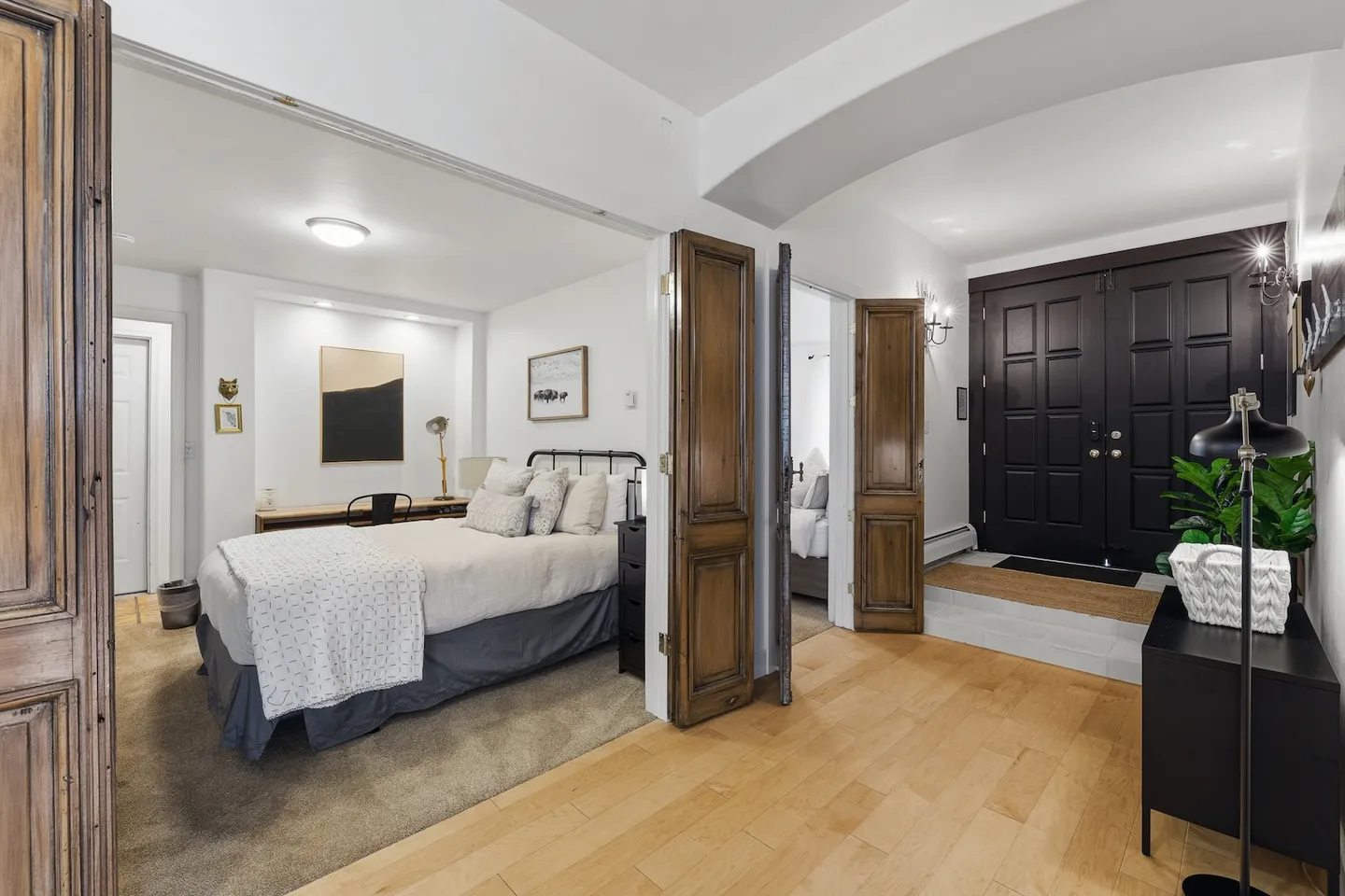 Entryway with black double front doors leading to light wood floors, with open double doors showing a bedroom with a bed and desk.